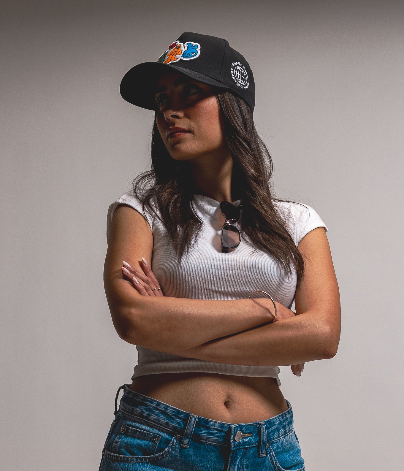 Woman wearing a white top, blue jeans, and a black cap with a logo on a gray background